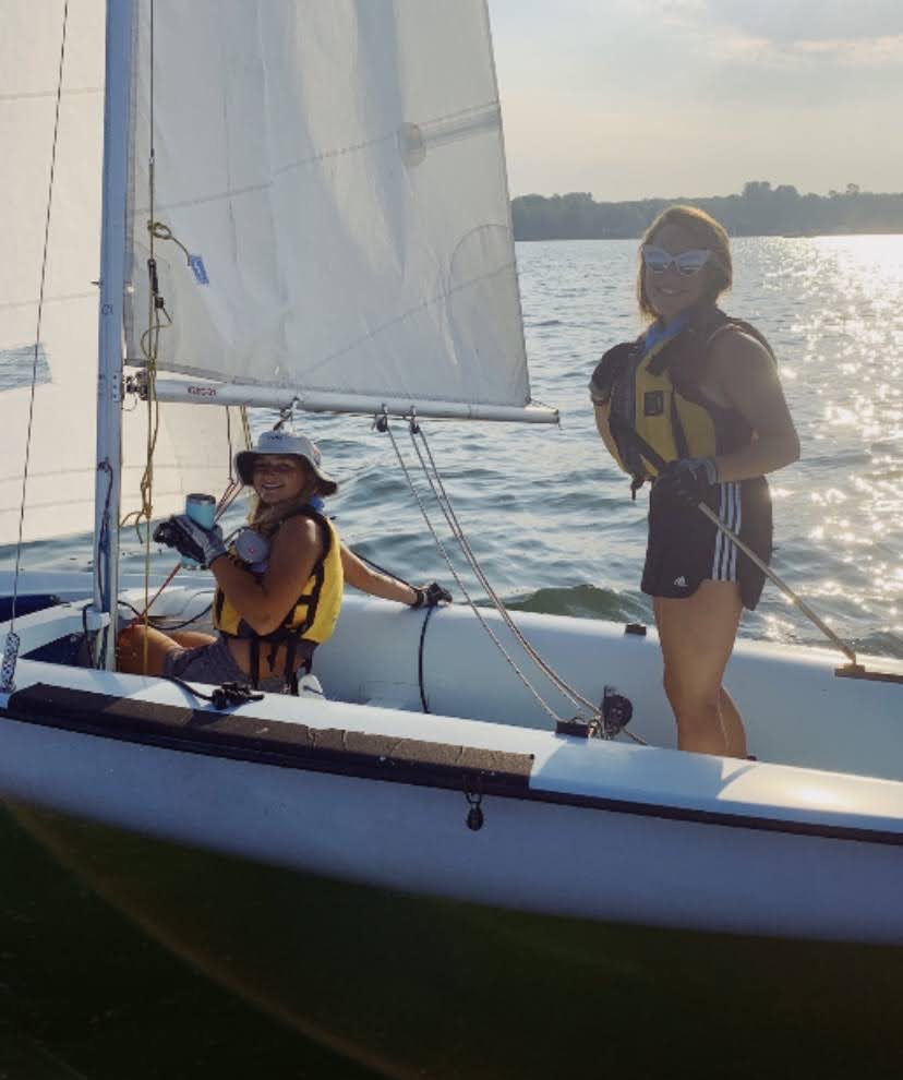 two people sailing a 420 sailboat one person is standing and one person is sitting they are sailing on a lake with a sunny day