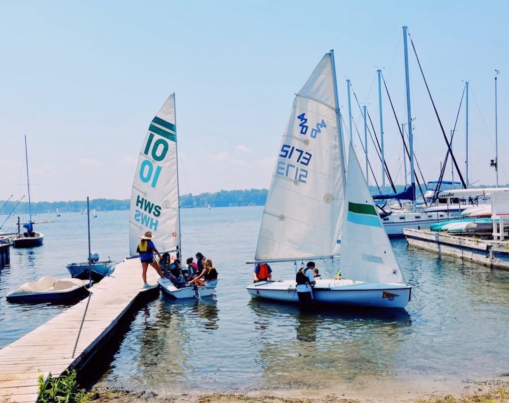 Two sailboats on the water with people getting the sailboats ready to launch